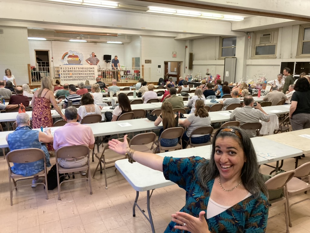 A large group of people playing bingo. A woman stands in front presenting the crowd.